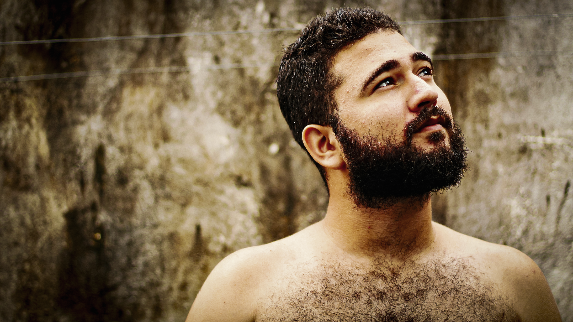 Portrait of a bearded man with a bare upper body looking upward in front of a textured concrete wall. The image is warmly and brightly lit, with a calm, natural and contemplative mood.