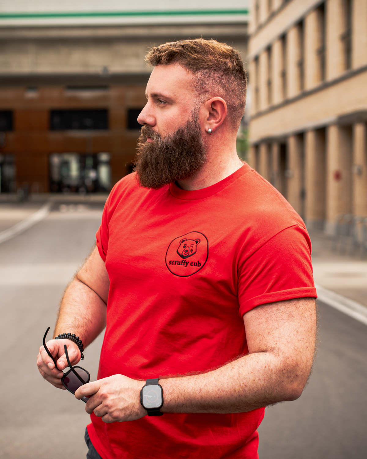 Bearded man wearing a red Scruffy Cub Anchor Collection bear T-shirt with embroidered logo, photographed outdoors.