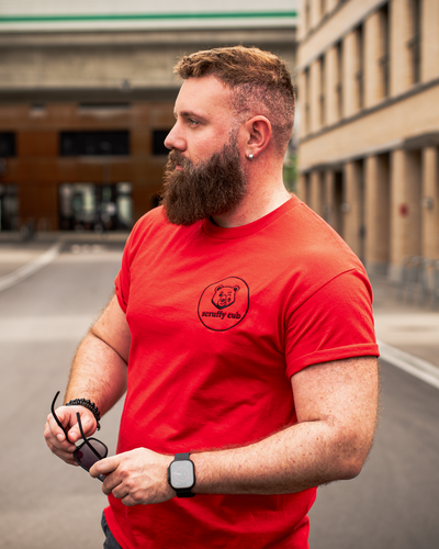 Bearded man wearing a red Scruffy Cub Anchor Collection bear T-shirt with embroidered logo, photographed outdoors.
