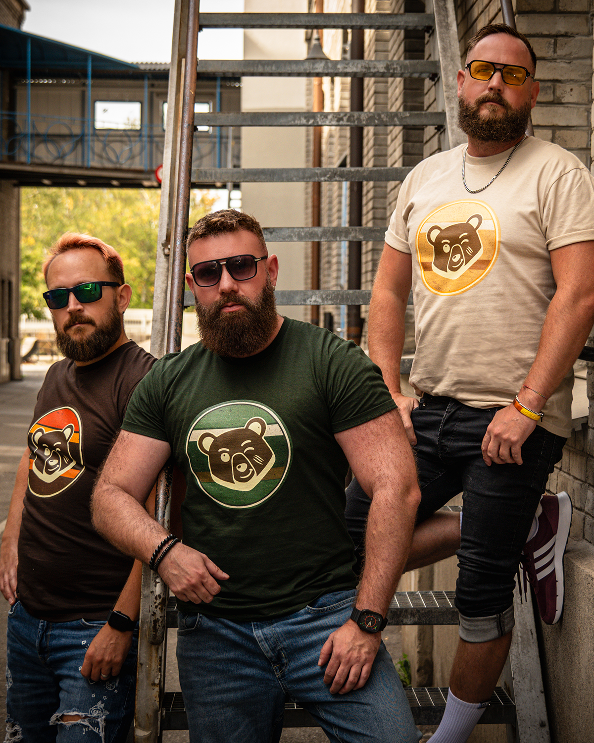 Three bearded men wearing Scruffy Cub Horizon T-shirts in sunset brown, desert beige and forest green - next to metal stairs