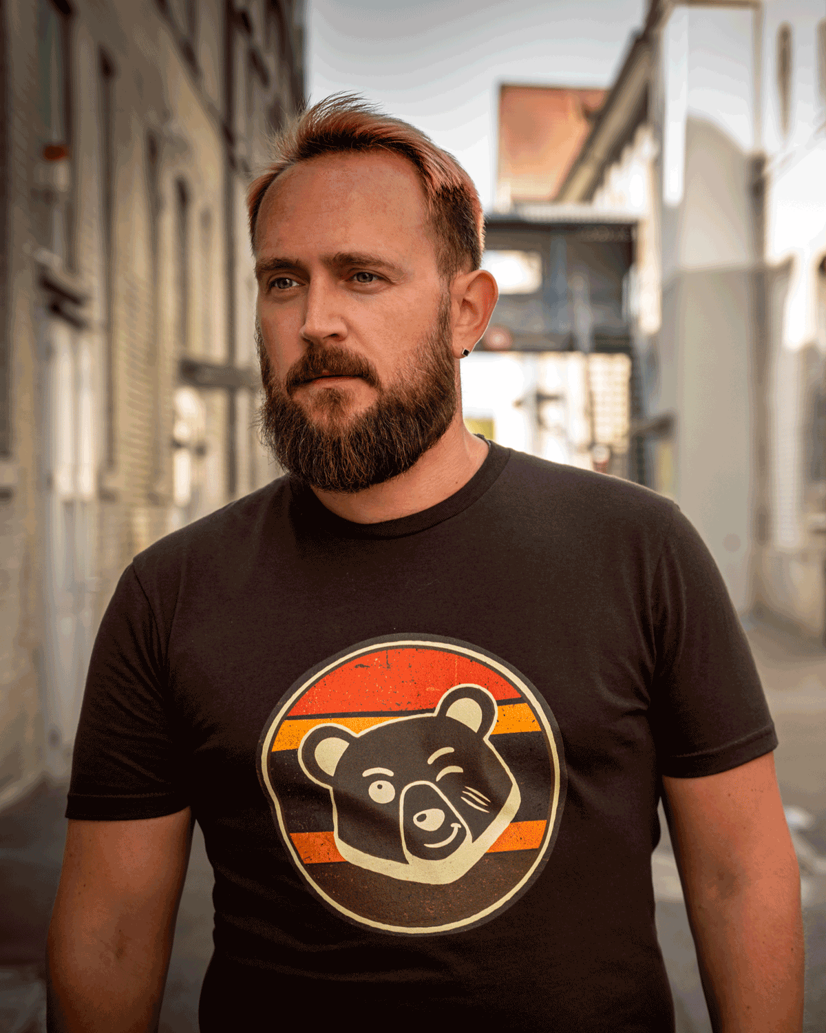 Bearded man wearing the sunset brown Scruffy Cub Horizon bear graphic T-shirt in an industrial alley