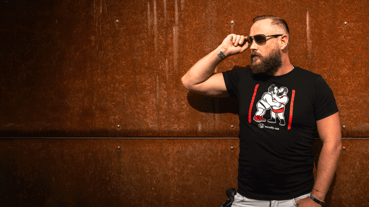 Bearded bearish man wearing a black Scruffy Cub Scratch Collection bear T-shirt with graphic print, posing in sunglasses against a rusted metal wall.