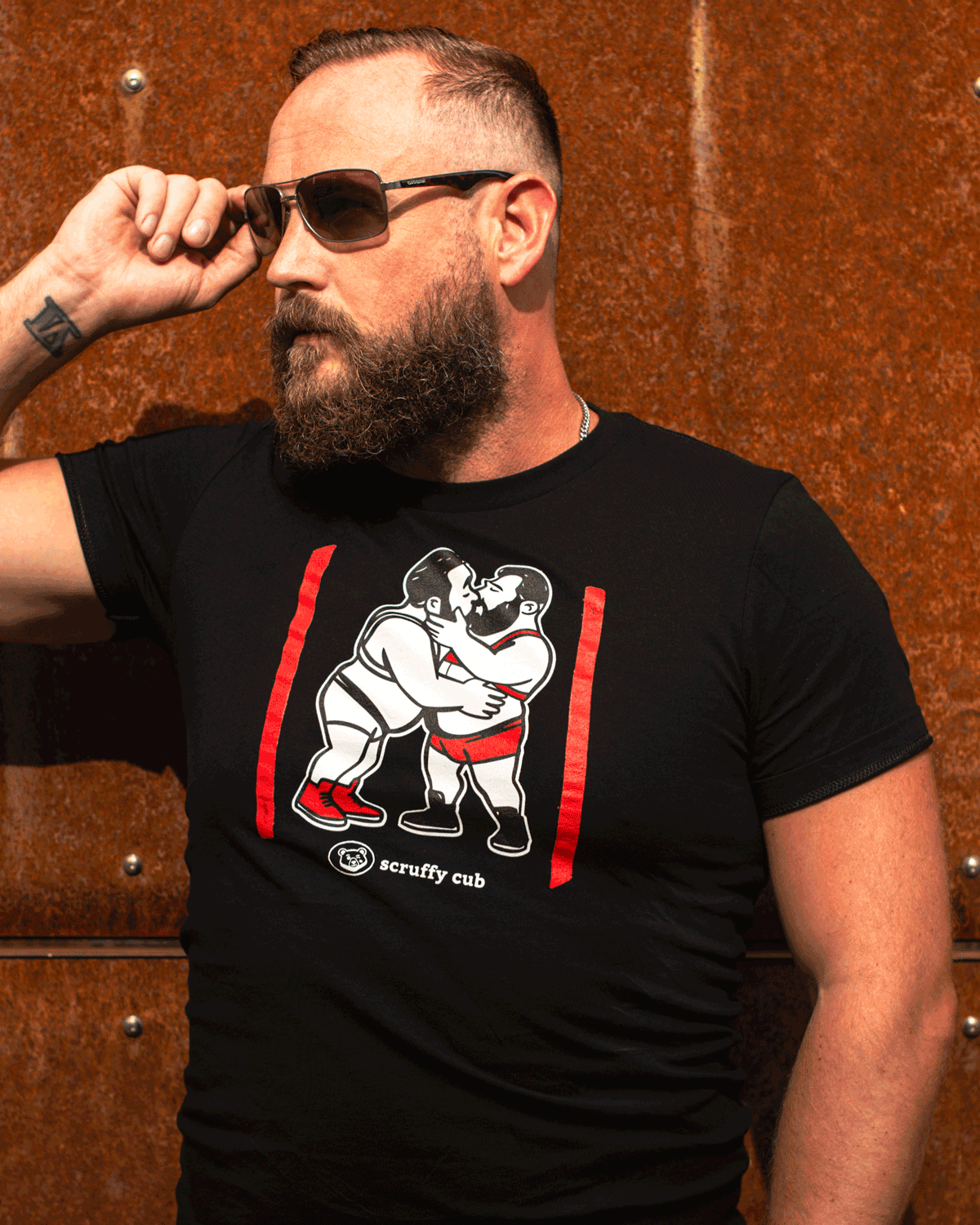 Bearded bear wearing a black Scruffy Cub Scratch Collection T-shirt with graphic print, posing in sunglasses against a rusted metal wall.
