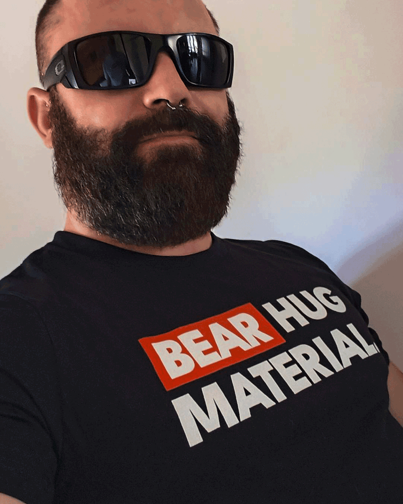 Bearded man wearing black sunglasses and a septum piercing, rocking the black Scruffy Cub “BEAR HUG MATERIAL” T-shirt with a bold red and white graphic print.